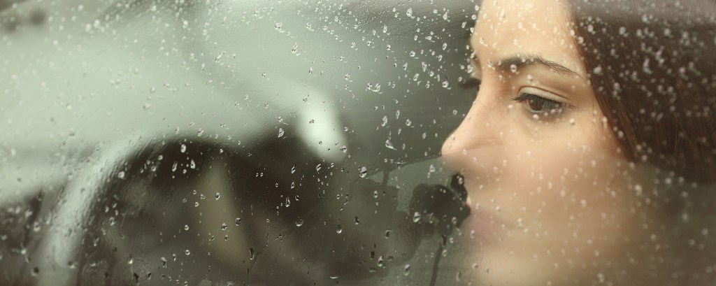 Mujer sentada en su carro durante una mañana lluviosa. No esta manejando. Esta observando a alguien que esta lejos.